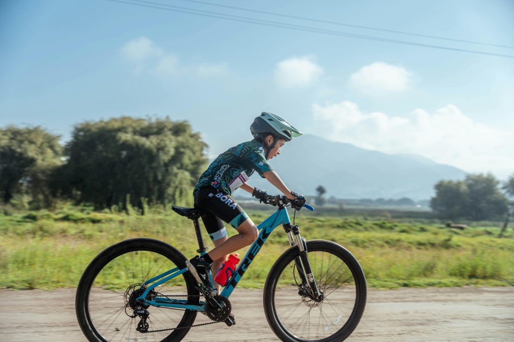 pexels-photo-17979478-17979478 A young boy in sportswear rides a mountain bike on a rural dirt road during a sunny summer day.